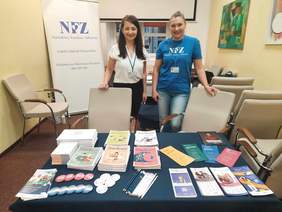 Pokaż zdjęcie: W pokoju stoją dwie panie. Uśmiechają się serdecznie. Przed nimi znajduje się stół, na którym leżą różne poradniki na temat zdrowia, ulotki z informacjami o programach zdrowotnych, gadżety reklamowe, ołówki. 