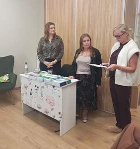 Pokaż zdjęcie: Trzy kobiety stoją przy stole z materiałami informacyjnymi i dokumentami. W tle znajduje się drewniana zabudowa, a na stole rozłożone są broszury, publikacje i napoje.