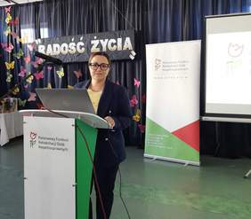 Pokaż zdjęcie: Przy mównicy z logo PFRON stoi kobieta ubrana w ciemny garnitur, w tle stoi rollup z logo funduszu. 