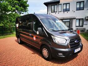 Pokaż zdjęcie: Srebrny bus stojący na parkingu.W tle biały budynek i drzewa