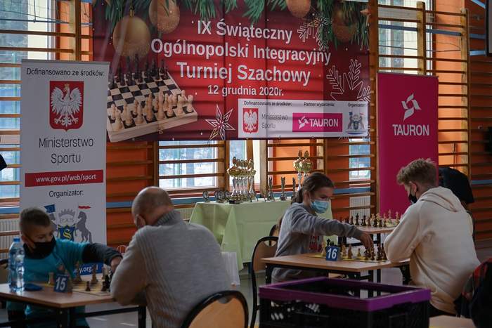 Pokaż zdjęcie: 2 stoliki przy których siedzą zawodnicy i grają w szachy w tle po bokach stoją podłużne banery ministerstwa sportu i firmy tauron na środku rozwieszony duży baner z napisem dziewiąty świąteczny ogólnopolski integracyjny turniej szachowy