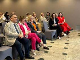 Pokaż zdjęcie: Grupa elegancko ubranych osób siedzi w sali konferencyjnej, uśmiechając się i słuchając prelekcji.