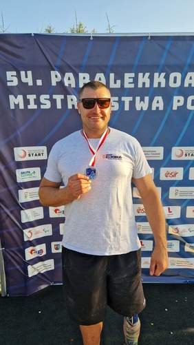 Pokaż zdjęcie: Zawodnik z medalem w ręku pozuje do zdjęcia na tle ścianki z napisem 54. Paralekkoatletyczne Mistrzostwa Polski