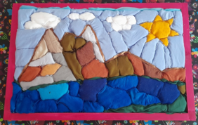 Pokaż zdjęcie: Zdjęcie: Praca z kategorii tkanina i aplikacja, na podkładzie formatu A3 stworzono kompozycję z kolorowej tkaniny, przedstawiającą jezioro w kilku odcieniach niebieskiego oraz góry wyłaniające się nad jeziorem. Po prawej stronie nad górami świeci żółte słońce.