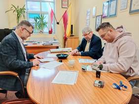 Pokaż zdjęcie: Przy podłużnym stole siedzi trzech mężczyzn. Podpisują umowę. Na stole leżą różne dokumenty, pieczątki, długopisy, telefony. 