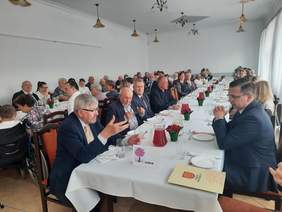 Pokaż zdjęcie: W sali stoją dwa rzędy stołów. Siedzą przy nich ludzie. Rozmawiają ze sobą i uśmiechają się do siebie. 