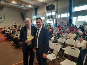 Pokaż zdjęcie: W sali gimnastycznej siedzą na krzesełkach ludzie. Oglądają występy artystyczne. Na pierwszym planie znajduje się dwóch mężczyzn w garniturach, którzy trzymają w dłoniach kwiaty. 