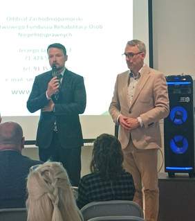 Pokaż zdjęcie: Dwóch wysokich mężczyzn ubranych w eleganckie garnitury. Pol lewej stronie Syrektor Oddziału PFRON w ciemnym garniturze z mikrofonem w ręku wita zgromadzonych gości, a po prawej Burmistrz Dębna w jasnym garniturze towarzyszy przy powitaniu. W tle panów ekran z prezentacją dotyczącą spotkania, a po prawej stronie w głębi podświetlony kolorowymi światłami głośnik.