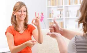 Pokaż zdjęcie: Naprzeciwko siebie siedzą dwie kobiety. Wykonują gesty w języku migowym. W tle widać regał z książkami i różowy kwiat.  