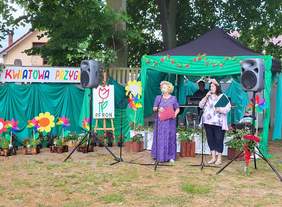 Pokaż zdjęcie: Prowadzące przy mikrofonie ubrane w kolorowe sukienki i mające wianki z kwiatów na głowie