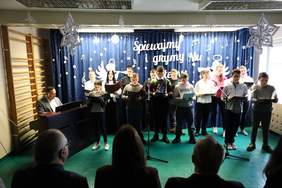 Pokaż zdjęcie: Grupa uczniów stojących na scenie śpiewa kolędy, część trzyma śpiewniki; za nimi dekoracja ze świątecznymi motywami, przed sceną widownia.