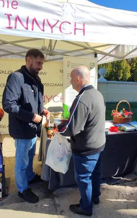 Pokaż zdjęcie: Mężczyzna przedstawiciel PFRON rozmawia z odwiedzającym podczas wydarzenia plenerowego. Na stole widoczne są broszury, ulotki i koszyk z materiałami promocyjnymi.