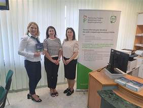 Pokaż zdjęcie: Trzy kobiety stojące obok siebie w jasnym pomieszczeniu biurowym. Po lewej stronie Ewa Siwik trzymająca folder informacyjny, obok niej Iwona Rogowska i Dorota Pieńkowska z PFRON. W tle roll-up z logo PFRON i informacjami o Centrach Informacyjno-Doradczych dla Osób z Niepełnosprawnościami CIDON.