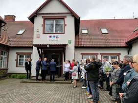 Na tle budynku WTZ Sławkowo uroczystość przecięcia wstęgi, a obok stoją uczestnicy obchodów otwarcia warsztatów