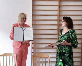 Pokaż zdjęcie: Dyrektor Marta Mordarska z uśmiechem patrzy na prezes Teresę Jaros-Ligaszewską, trzyma list gratulacyjny, który otrzymała od Stowarzyszenia