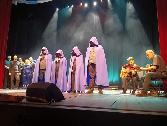 Pokaż zdjęcie: Scena, a na niej grupa czterech artystów ubranych w fioletowe płaszcze z kapturami. Obok siedzi akompaniująca para. 