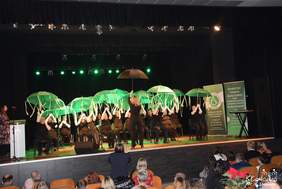 Pokaż zdjęcie: Na scenie w rzędzie siedzi grupa osób, trzymając nad głowami zielone parasole. Czarne tło podświetlają zielone reflektory. Przed sceną znajduje się widownia z uczestnikami. Na scenie stoją dwa pionowe banery Warsztatu Terapii Zajęciowej w Kłodawie  oraz Stowarzyszeniu „Sprawni Inaczej” w Kłodawie