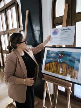 Pokaż zdjęcie: Kobieta w beżowej marynarce poprawia informację o wystawie zamontowaną na sztaludze z pracą plastyczną.