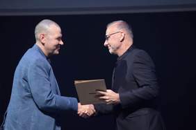 Pokaż zdjęcie: Dwaj mężczyźni w marynarkach ściskają sobie dłonie, jeden z nich w okularach trzyma teczkę z napisem Gdynia.