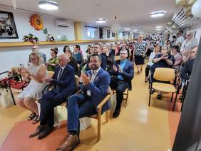 Pokaż zdjęcie: Na krzesełkach siedzą zaproszeni goście, ubrani w garnitury i eleganckie sukienki.
