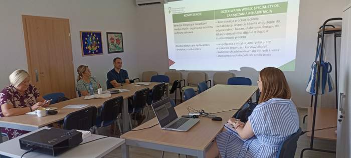 Pokaż zdjęcie: Widoczna prezentacja, przy stolikach siedzą trzy kobiety i jeden mężczyzna