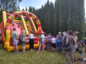 Pokaż zdjęcie: Dwóch chłopców zjeżdża z zjeżdżalni na brzuchu z głową do dołu. Przy zjeżdżalni tworzy się kolejka dzieci z rodzicami. 