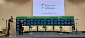 Pokaż zdjęcie: Na scenie przy mównicy stoi Pan Krzysztof Czechowski Koordynator ds. dostępności ZUS oraz Pani Anna Wandzel Dyrektor Oddziału Mazowieckiego PFRON, w tle slajd prezentacji przedstawiający logo PFRON.