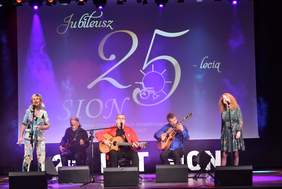 Pokaż zdjęcie: Na scenie występuje pięcioosobowy zesół. Po bokach śpiewają dwie wokalistki, w środku trzech siedzących gitarzystów,