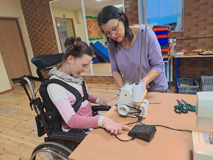 Pokaż zdjęcie: Dziewczynka na wózku, przy pomocy opiekunki, uczy się szyć na maszynie i jest bardzo uśmiechnięta i zadowolona
