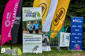 Pokaż zdjęcie: Zbliżenie na banery i flagi promujące 5 km Bieg Pogoń Raka, który odbywa się "zawsze w pierwszą sobotę października". Na jednym z banerów widać uśmiechniętych biegaczy w niebieskich koszulkach z numerami. Widoczne są też loga sponsorów i partnerów.