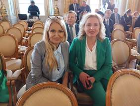 Pokaż zdjęcie: Zdjęcie dwóch kobiet blondynek, siedzących na krzesłach, pozują do pamiątkowego zdjęcia.