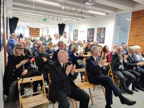 Pokaż zdjęcie: Zdjęcie sali i zaproszonych gości podczas uroczystości jubileuszowych. Goście siedzą na krzesłach, elegancko ubrani, spoglądają w kierunku sceny, część  osób klaszcze. 