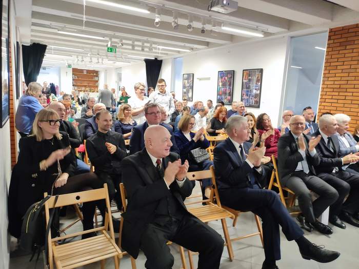 Pokaż zdjęcie: Zdjęcie sali i zaproszonych gości podczas uroczystości jubileuszowych. Goście siedzą na krzesłach, elegancko ubrani, spoglądają w kierunku sceny, część  osób klaszcze. 