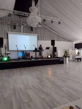 Kobieta na scenie, widok z dalszej perspektywy, stoi za mikrofonem, wystrój sali i kolor podłogi chłodny beż