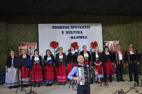 Pokaż zdjęcie: Na scenie stoją kobiety i mężczyźni przebrani w stroje ludowe, przed nimi stoi mężczyzna z harmonią na której gra