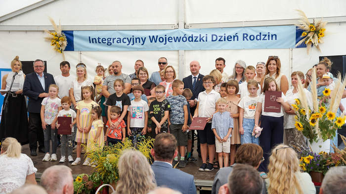 Pokaż zdjęcie: Pamiątkowe zdjęcie rodzin adopcyjnych z dziećmi. Grupa osób z dyplomami stoi na scenie. 