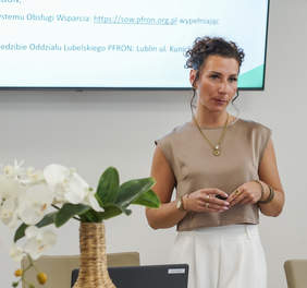 Pokaż zdjęcie: Prelegentka młoda kobieta stoi przy stole konferencyjnym, w tle na ścianie slajd z prezentacją.