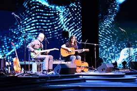 Fotografia przedstawia scenę, oświetloną niebieski światłem. Na scenie siedzą dwie osoby z gitarami. Kobieta gra na gitarze akustycznej, mężczyzna gra na elektrycznej. Przed nimi na scenie znajdują się elementy nagłośnienia (głośniki)