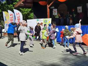 Pokaż zdjęcie: Na chodniku tańczą dzieci. Są ubrane na kolorowo. W tle widać scenę i parkowe drzewa. 