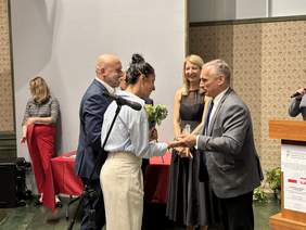 Pokaż zdjęcie: Mężczyzna w marynarce koszuli i krawacie przekazuje szklaną statuetkę w kształcie skały kobiecie w ciemnych krótko spiętych włosach koszuli w prążki i spodniach. Za nimi uśmiecha się kobieta w blond włosach i w czarnej sukni.