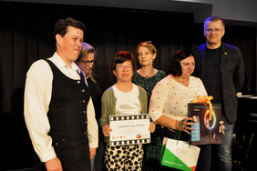 Pokaż zdjęcie: Dwóch mężczyzn oraz cztery kobiety stoją na scenie. Jedna z kobiet trzyma w dłoniach dyplom, natomiast druga trzyma nagrodę rzeczową oraz torbę z logo PFRON.