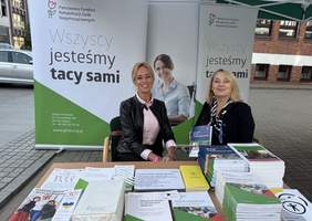 Pokaż zdjęcie: Za biurkiem siedzą dwie uśmiechnięte kobiety ubrane w marynarki. Na stole leży dużo materiałów informacyjnych z logo PFRON. Za nimi stoi duża ścianka zrobiona z 3 banerów z logo PFRON.