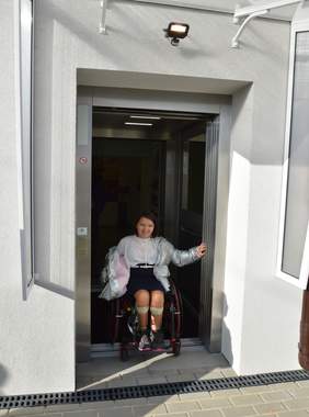 Pokaż zdjęcie: na zdjęciu budynek szkoły w której znajduje się winda zewnętrzna z której wyjeżdza dziewczynka poruszająca się na wózku inwalidzkim