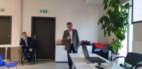 Sala szkoleniowa. Pan Łukasz Żmuda - Dyrektor Oddziału Opolskiego PFRON stoi przed ekranem, na którym wyświetlana jest prezentacja. Odwrócony w stronę uczestników spotkania opowiada o programach realizowanych przez PFRON