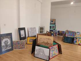 Pokaż zdjęcie: Fragment dużego pomieszczenia na tle białych ścian ustawione są obrazy 