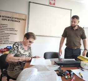 Pokaż zdjęcie: Mężczyzna stoi obok kobiety, która siedzi przy biurku. Kobieta czyta. Znajdują się w pomieszczeniu, za nimi baner GOPS oraz tablica.