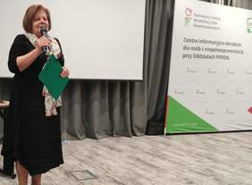 Kobieta w czarnej sukience i kolorowym szalu na szyi, z mikrofonem w ręku, stoi