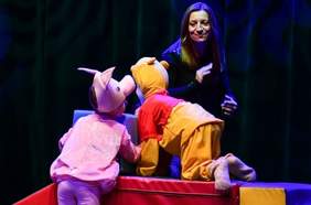 Pokaż zdjęcie: Na scenie na kolorowych klockach bawią się małe dzieci przbrane za kubusia puchatka i prosiaczka, z tyłu przygląda im się pani opiekunka