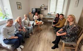 Pokaż zdjęcie: Grupa ośmiu osób siedzi w kręgu w przytulnym pokoju z oknem i stolikiem kawowym na środku.