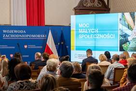 Pokaż zdjęcie: widok na gości spotkania siedzących na krzesłach od tyłu, w tle ekran z napissem „Moc Małych Społeczności” w Szczecinie, a po lewej stronie baner województwa zachodniopomorskiego